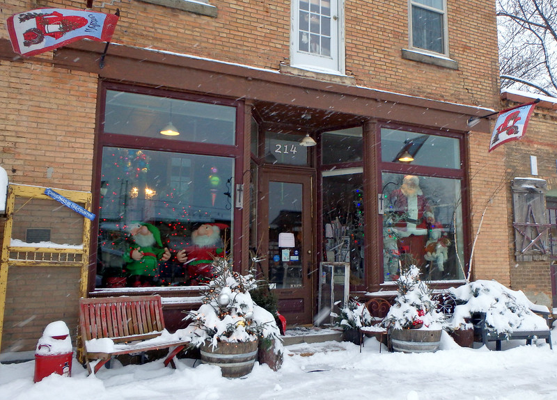 two large elves in one window, Santa in the other, snow falling and covering the decorated pots in front
