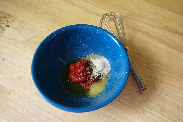 A blue bowl filled with unmixed sauce components: a mound of tomato sauce ringed with a small pool of olive oil, and herbs, garlic powder, and salt scattered on top