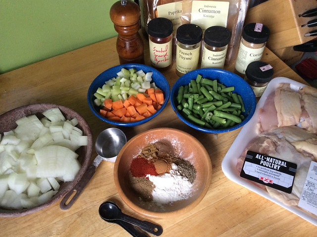 Bowl of chopped vegetables -- onions, carrots, celery, green beans -- sit on a counter alongside a bowl of flour spotted with small hillocks of spices, as well as a large styrofoam package of chicken thighs, all in front of a wall of spice jars and bags.