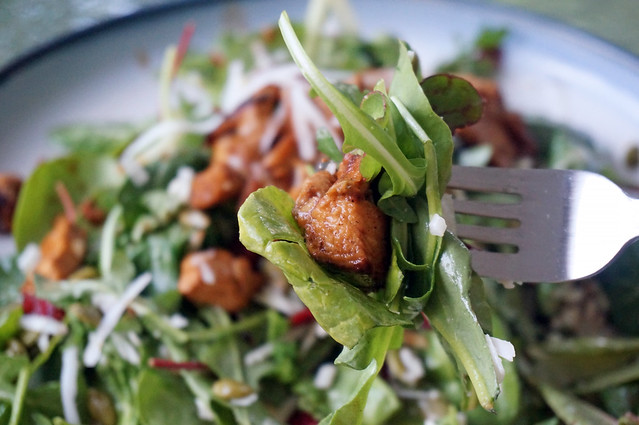 A bite salad speared on a fork, with the rest of the salad blurry in the background. One single chunk of chicken is half-wrapped in a large yellow-green leaf, other smaller leaves tangled around it, with shreds of cheese stuck to the various leaves.